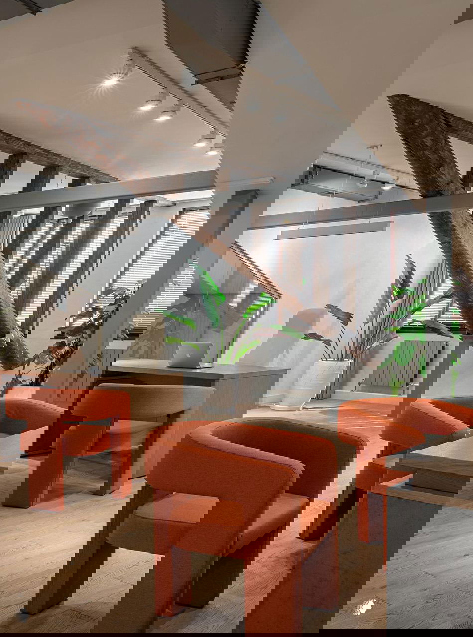 Помещение с деревянными балками и цилиндрическими оранжевыми креслами, растениями и дневным светом из окон — сочетание исторических элементов и современного дизайна.