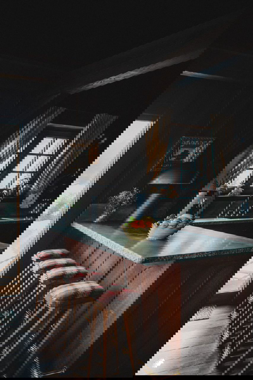 Кухонная зона в темных деревянных тонах: барная стойка с зелёной столешницей, деревянные барные стулья с клетчатой обивкой и окно в альпийском стиле.