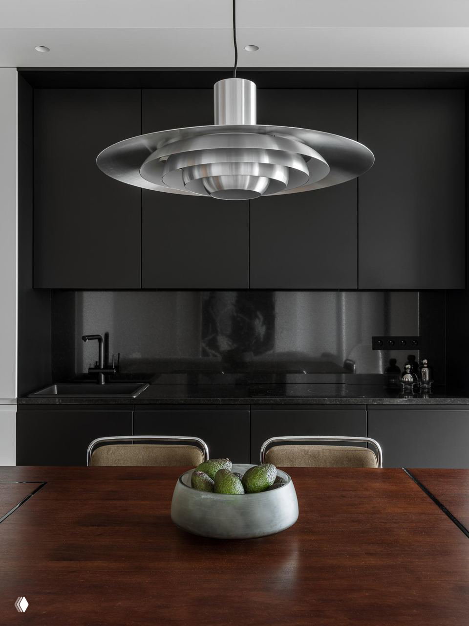 Крупный план столовой зоны: деревянная столешница, чаша с фруктами и массивная многослойная металлическая подвесная лампа над столом.