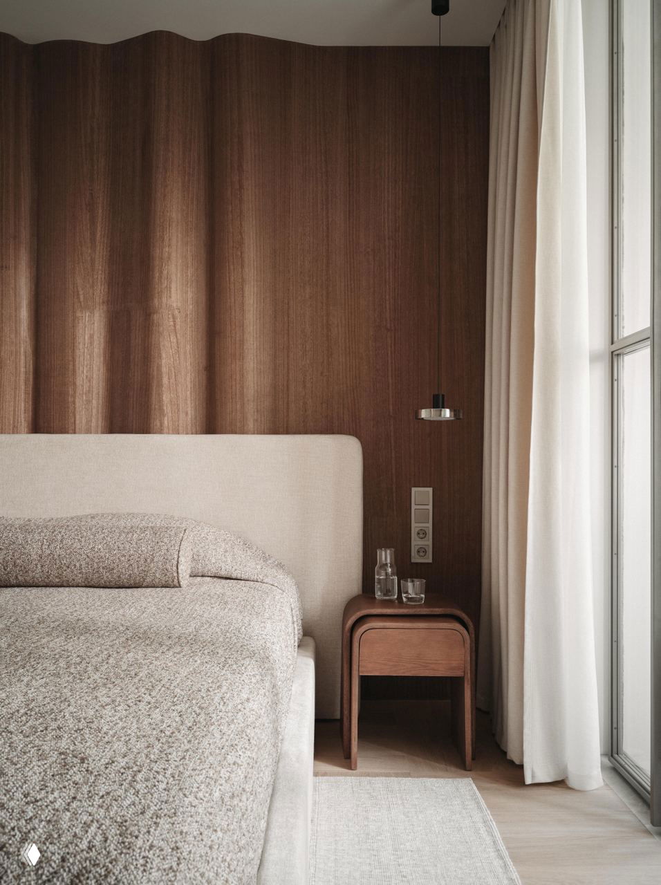 Спальня с волнообразной деревянной стеной за изголовьем, светлая обивка кровати и деревянная тумбочка в теплой коричневой гамме.
