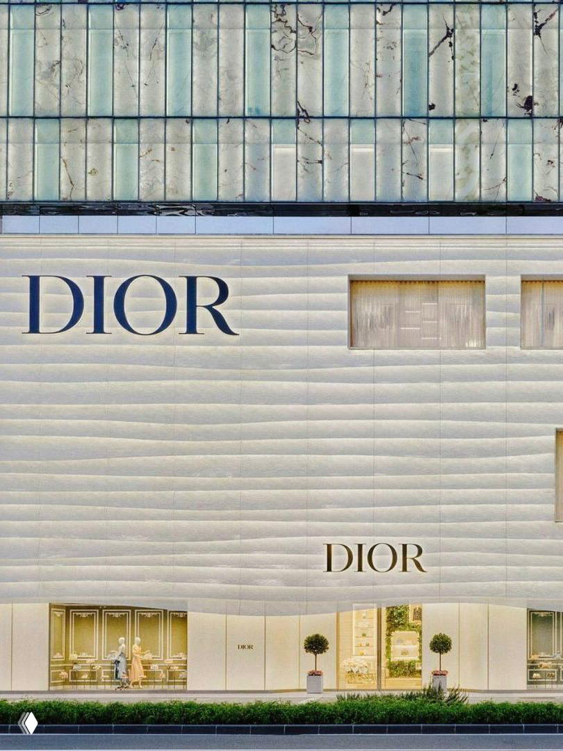 Крупный план фасада бутика Dior: волнообразная белая облицовка, витрины первого этажа, заметный логотип DIOR и аккуратные растения у входа.