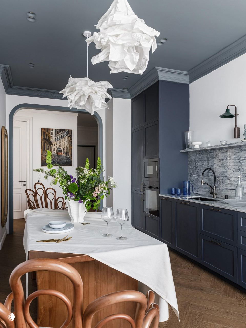 Кухня-столовая с тёмно-синим потолком и молочно-белыми стенами, крупная подвесная лампа и деревянная мебель создают ощущение уюта и камерности.