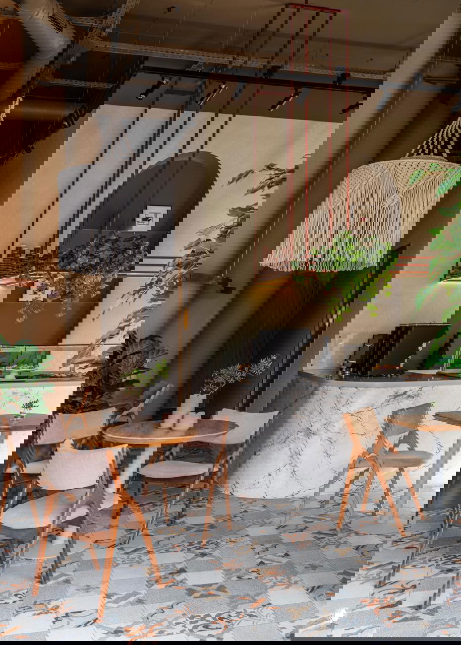 Общий вид кафе: макраме-люстра, круглые столы, деревянные стулья и мраморная стойка на фоне тёплых природных оттенков и растений.