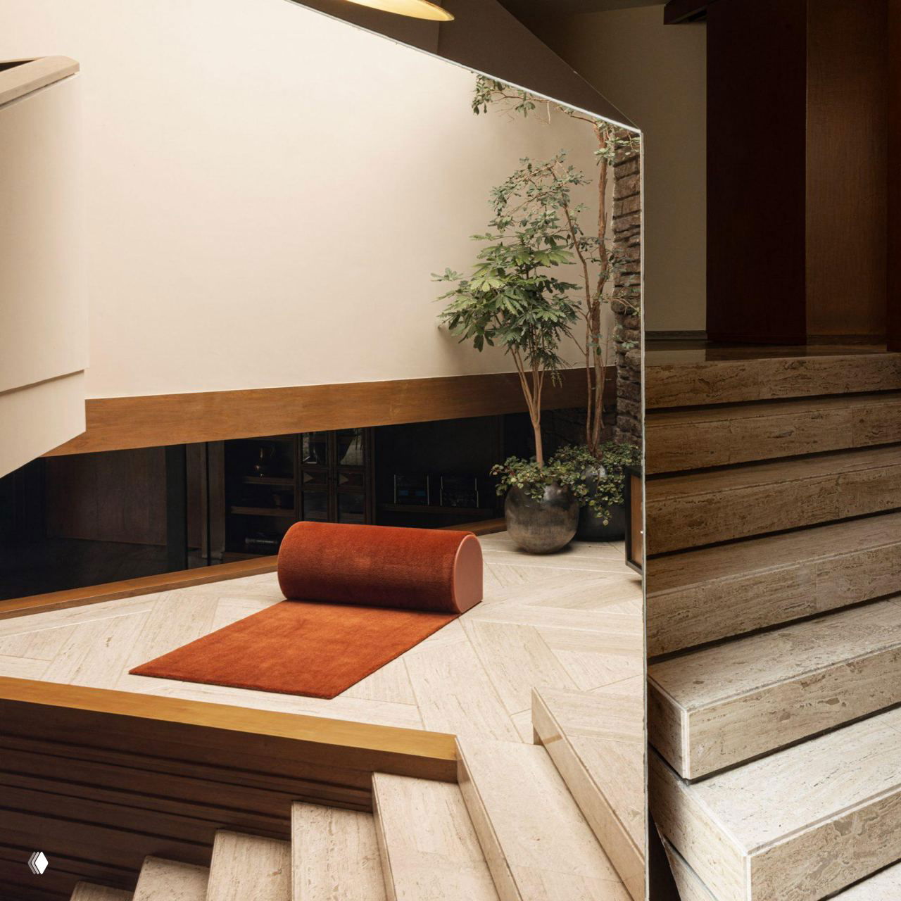 Ковер Roll в терракотовом цвете развёрнут на светлом каменном полу у лестницы: аккуратный валик у края и минималистичный интерьер в кадре.