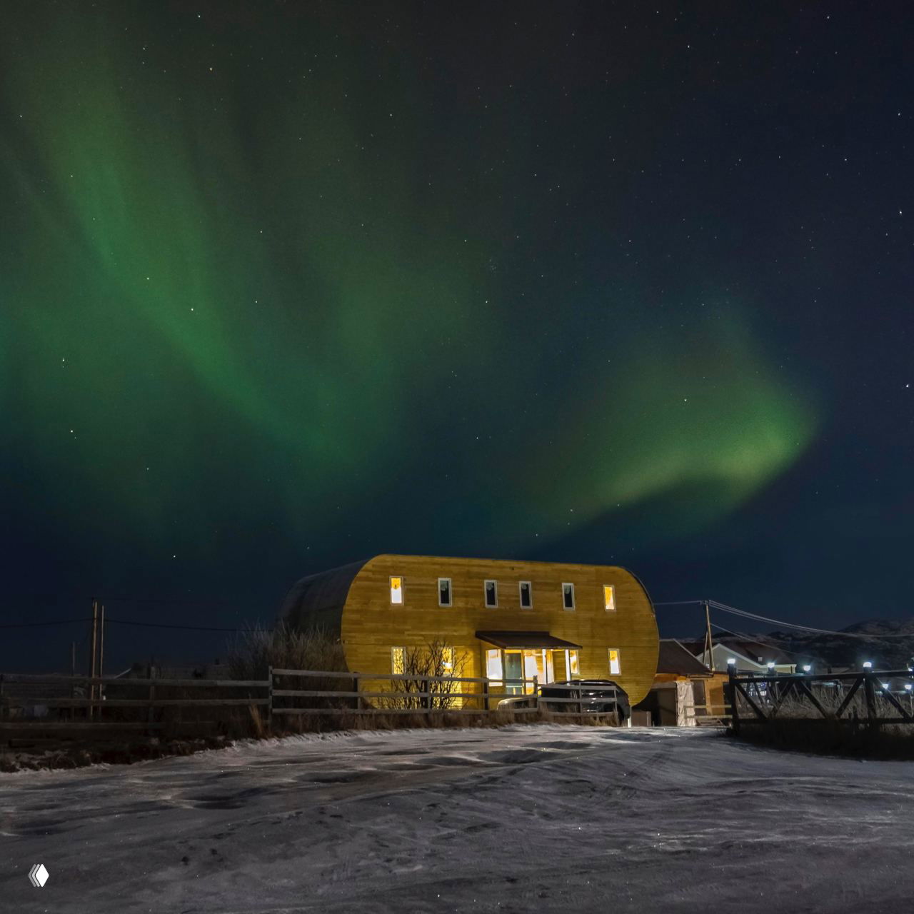 Ночной домик в Териберке под северным сиянием: тёплое освещение, снега вокруг и зелёные волны сияния над тёмным небом.