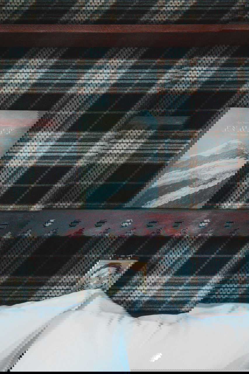 Крупный план книжной полки и клетчатой обивки в шале: книга с изображением гор и настольные часы на фоне тёмного древесного панно.