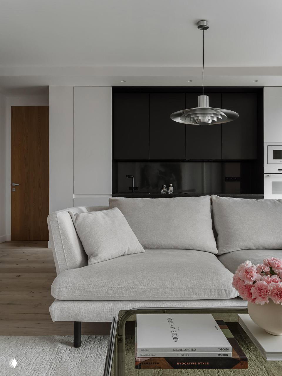Открытая планировка: светлый диван на фоне тёмной кухонной стены и подвесного светильника; минималистичная мебель и нейтральная гамма.