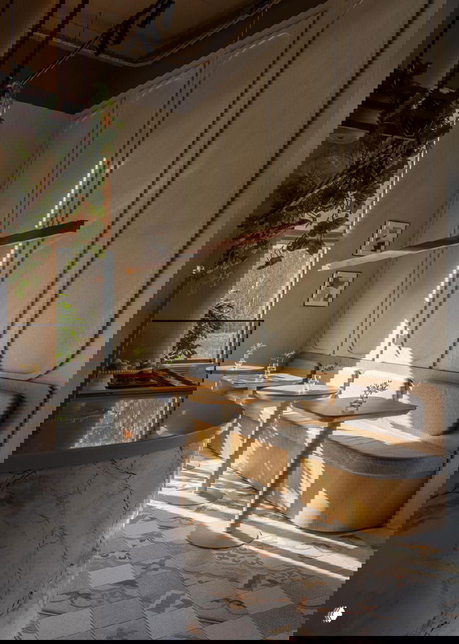 Уютная зона у окна: мягкая скамья, небольшие столики, высокие стеновые панели и рассеянный солнечный свет, создающий уютную атмосферу.