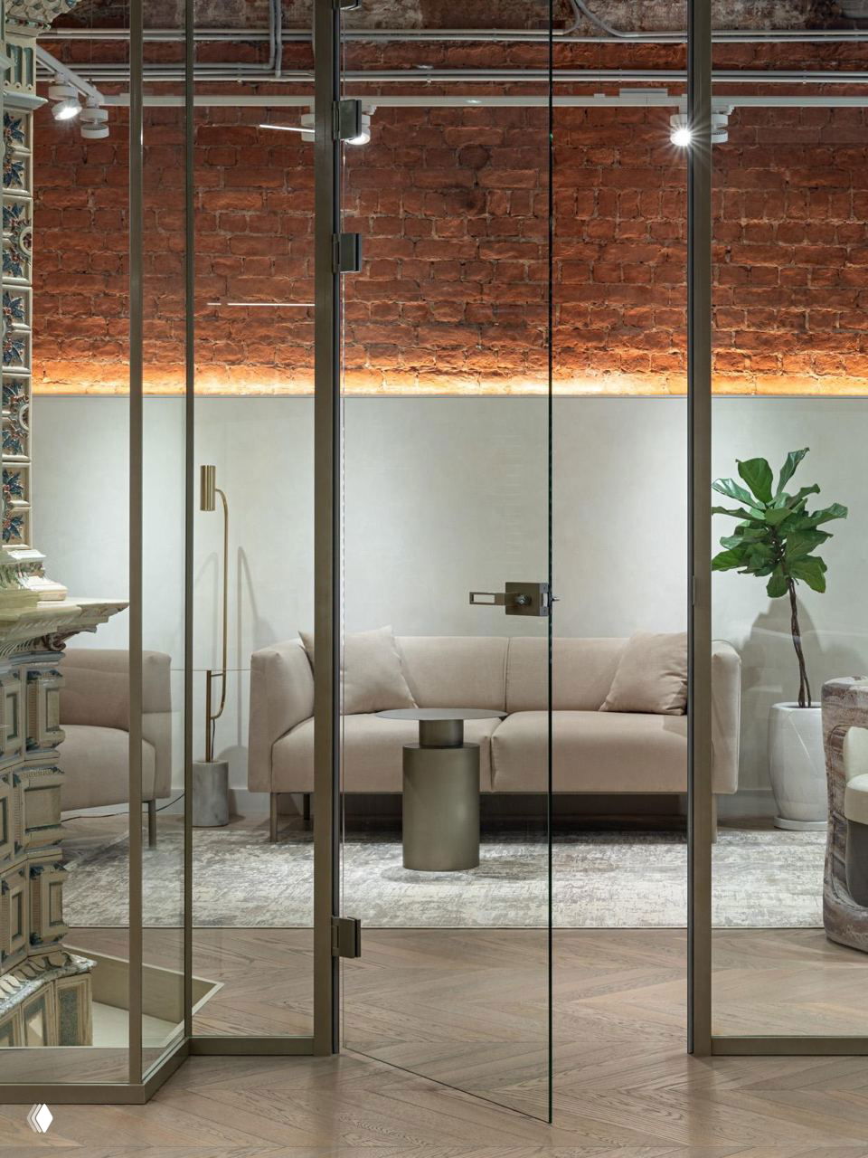 Зона отдыха за стеклянной дверью: бежевый диван, журнальный столик и мягкое освещение на фоне стены с декоративным кирпичом и растением.