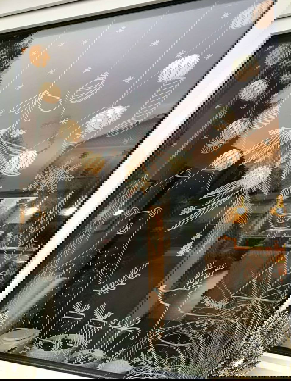 Окно с белыми контурными рисунками на стекле — домики, подарки, лесные мотивы и висящие гирлянды, гармонирующие с интерьерной подсветкой внутри.