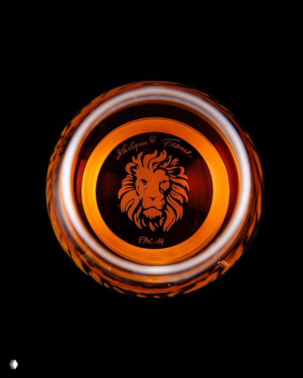 Вид снизу бутылки: выгравированная голова льва на дне стеклянного декора Lalique и тёплые янтарные оттенки коньяка.