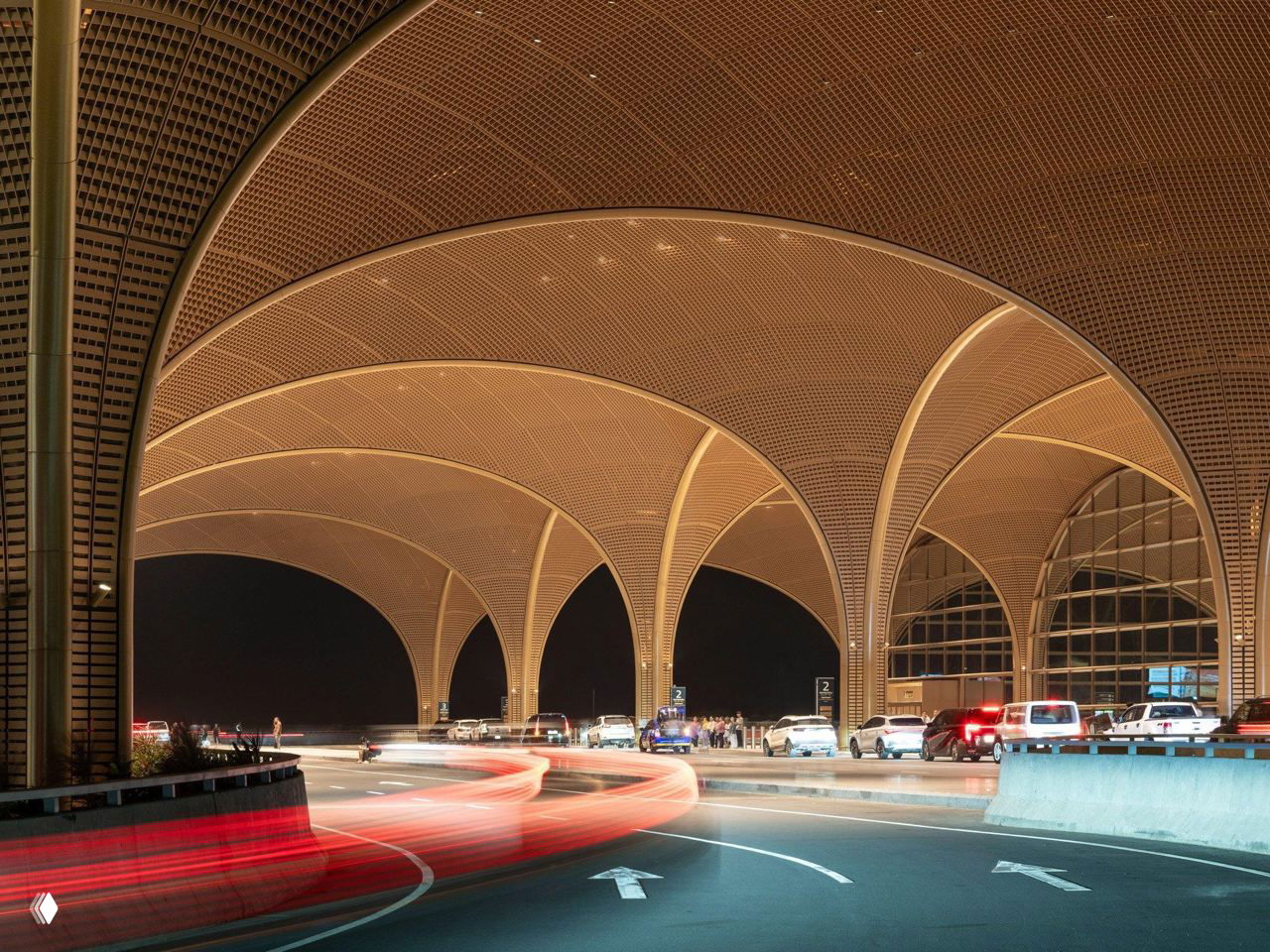 Ночной вид подъездной зоны аэропорта: подсвеченные арочные своды, навесы над подъездом и световые следы автомобилей в движении на дороге у терминала.