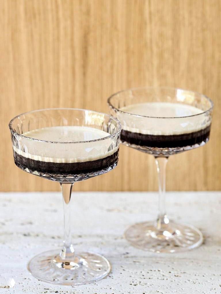 Два элегантных бокала купе с кремовой пеной эспрессо мартини на светлом фоне, минималистичная праздничная подача в паре бокалов.