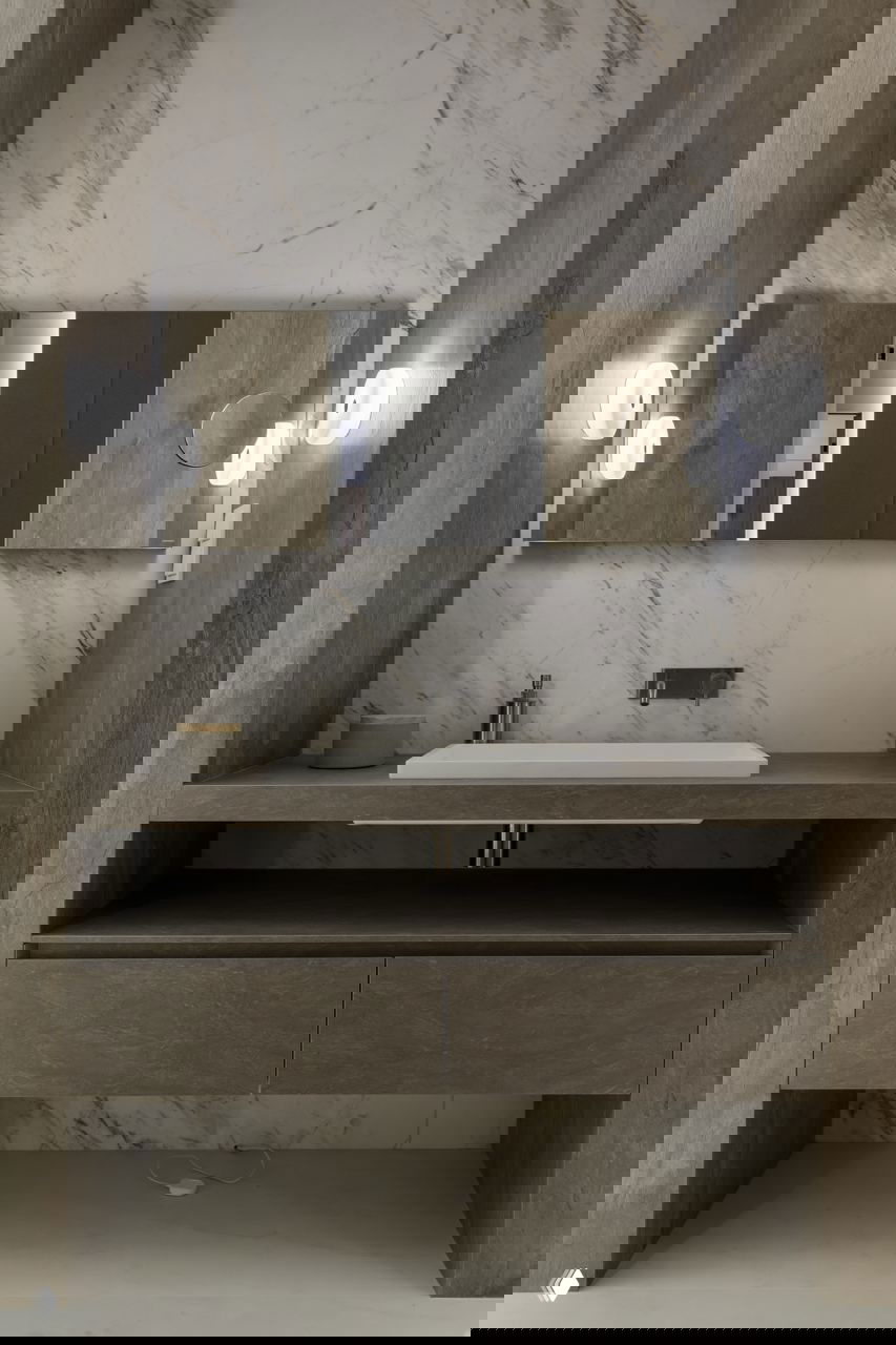 Ванная комната с мраморной облицовкой, встроенной тумбой под умывальник и крупным зеркалом, материалы и освещение подчёркивают текстуры и тональность интерьера.