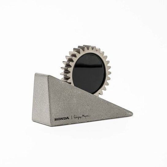 Компактная фоторамка-геар: металлическая зубчатая шестерня встроена в наклонный бетонный блок с зеркальной вставкой, студийная съемка на белом фоне.