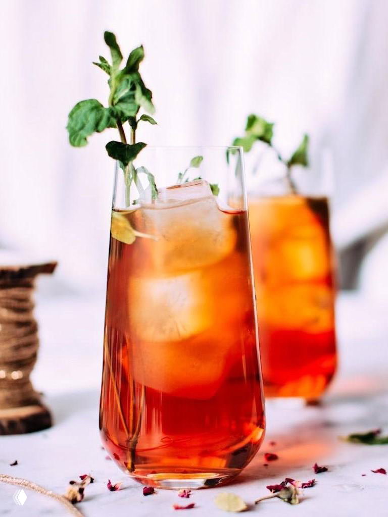 Два высоких стакана с янтарно-оранжевым ледяным коктейлем, кубики льда и зелень мяты как украшение, светлый праздничный фон.