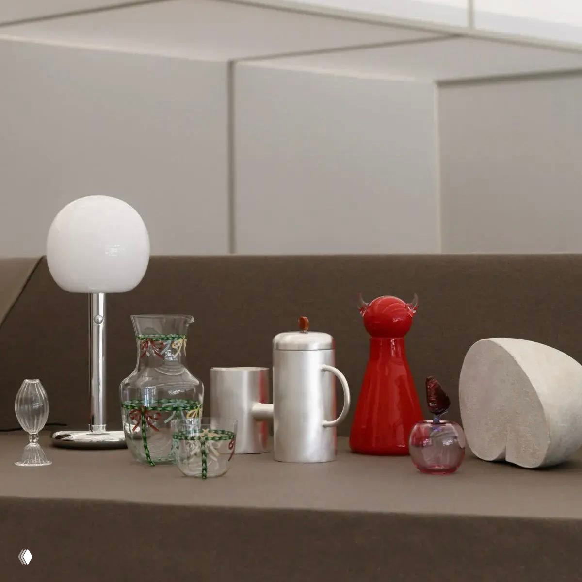 Натюрморт из предметов дизайна: настольная лампа, стеклянные графины, керамические сосуды и скульптурные формы на нейтральном фоне.