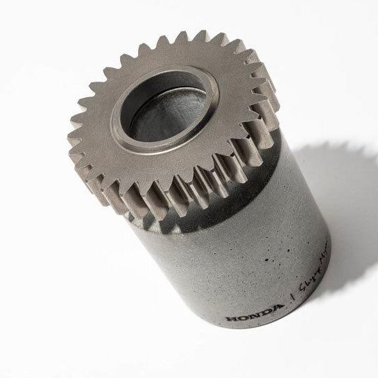 Крупный план зубчатой шестерни, погружённой в бетонный блок: текстура металла и бетона, отпечаток подписи Honda на основании, студийный свет.