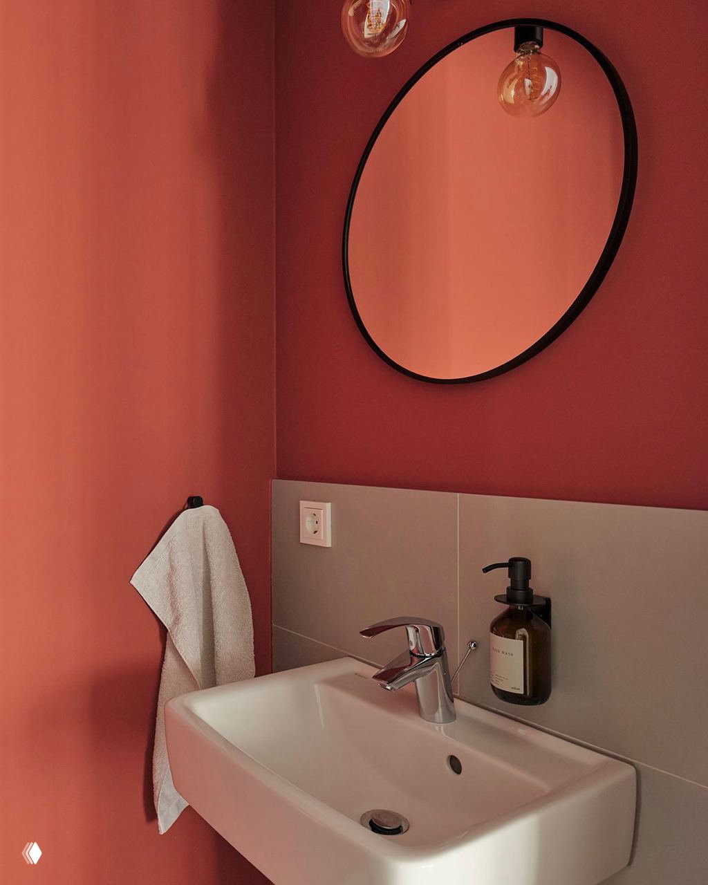 Ванная зона с насыщенной терракотовой стеной, круглым зеркалом в тон чёрных декоративных деталей и минималистичной белой раковиной, тёплый стиль.