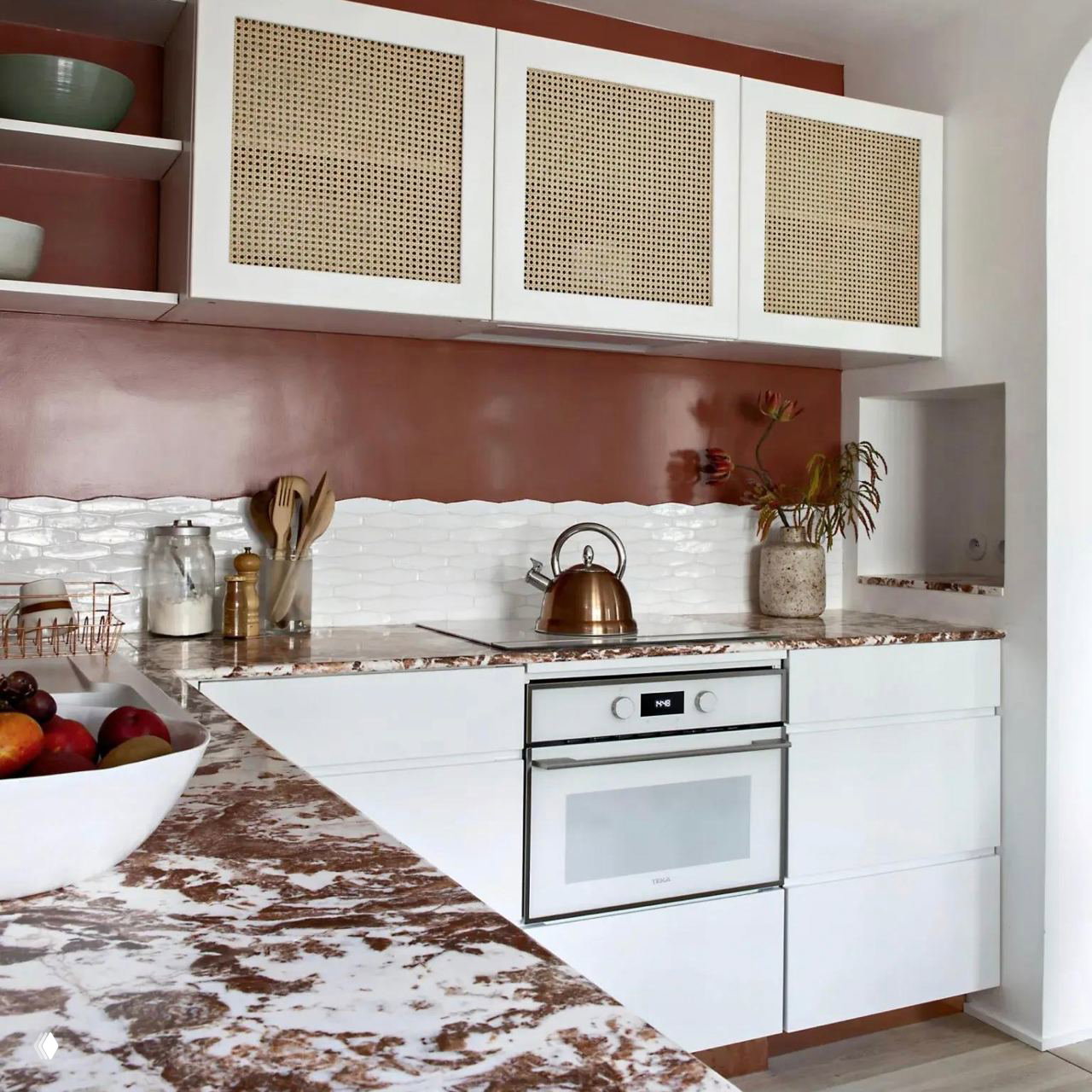Кухонный уголок с белыми фасадами, мраморной столешницей и тёплой розово-терракотовой стеной; встроенная ниша видна сбоку.