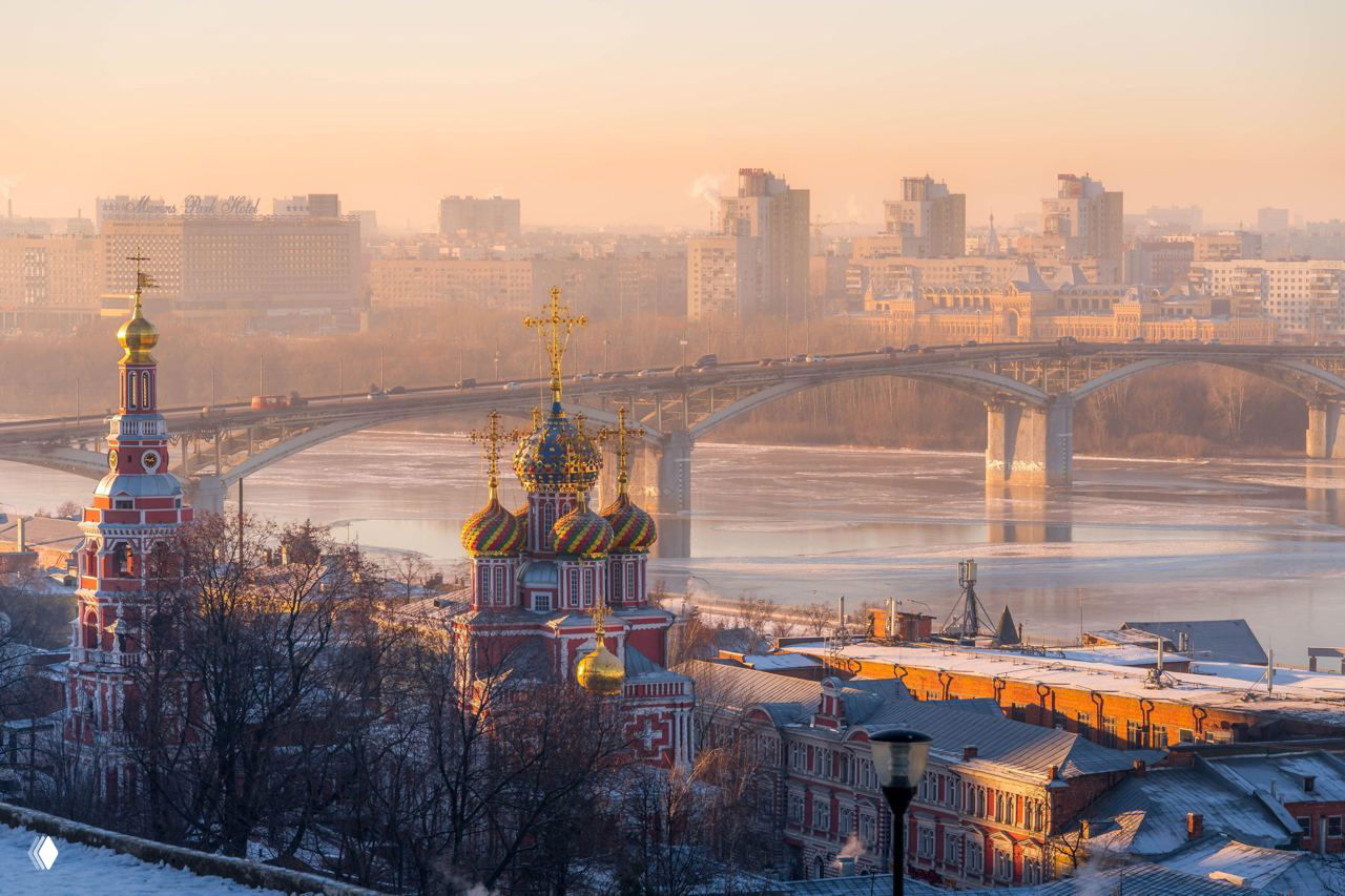 Панорамный вид на Нижний Новгород зимой: яркие купола храма, река Ока и мост в тёплом утреннем свете.