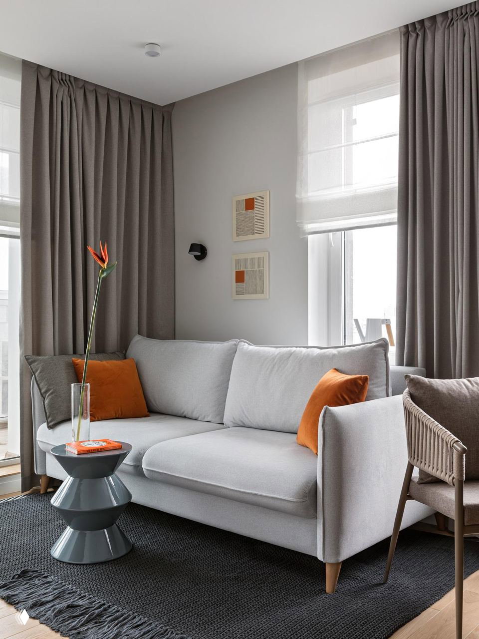 Гостиная с серым диваном и яркими оранжевыми декоративными подушками, высокие серые шторы и небольшая декоративная табуретка создают баланс цвета и формы