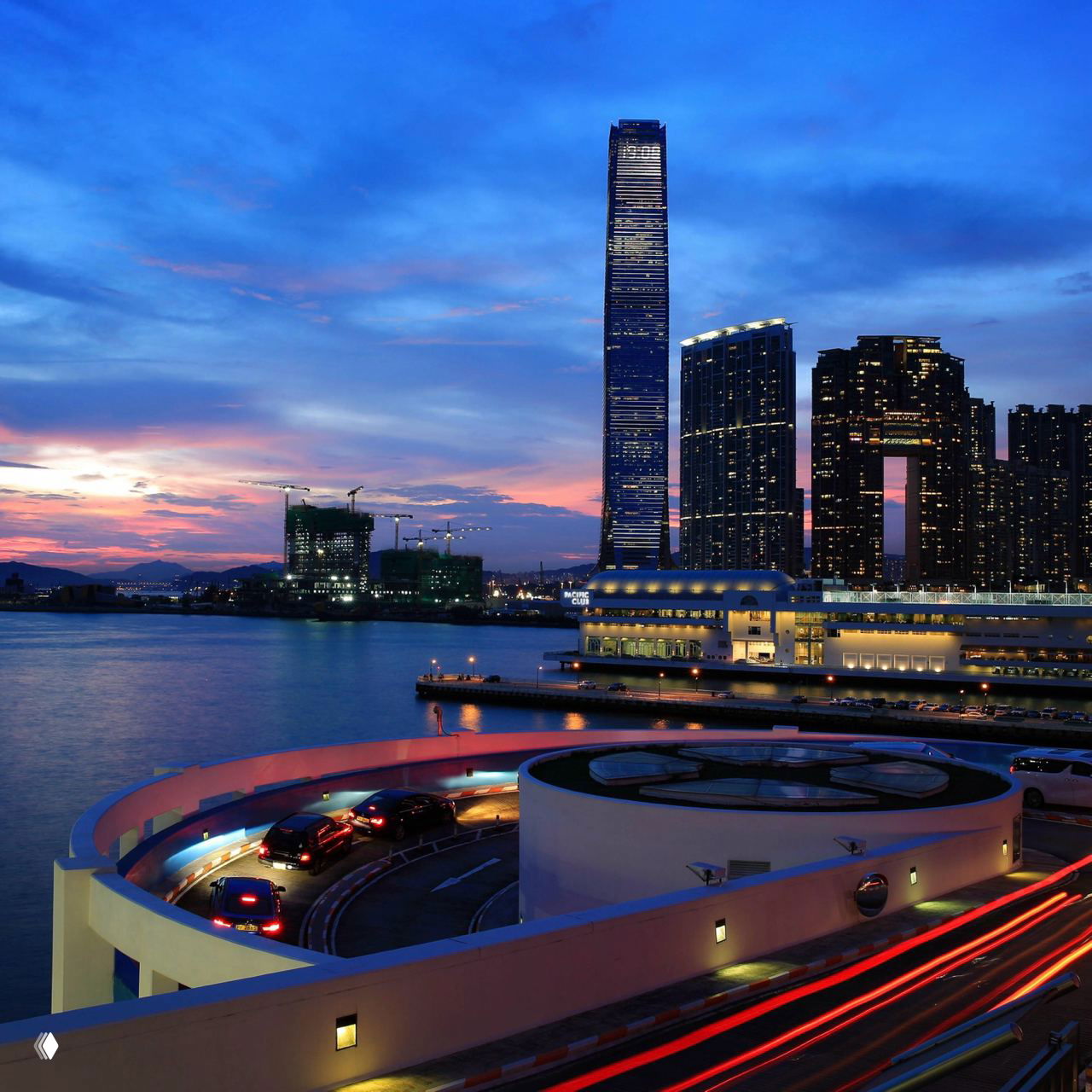 Горизонт Гонконга на закате: высотные башни у набережной и извилистая дорожная спираль на переднем плане в мягком сумеречном свете.