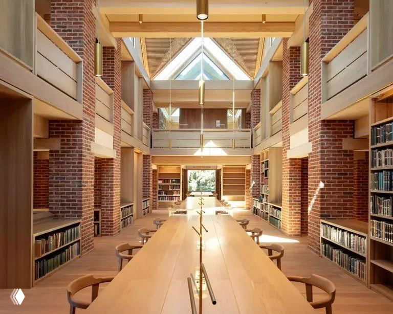 Интерьер новой библиотеки с длинным деревянным столом, стульями, кирпичными стенами и стеллажами для книг при дневном свете.