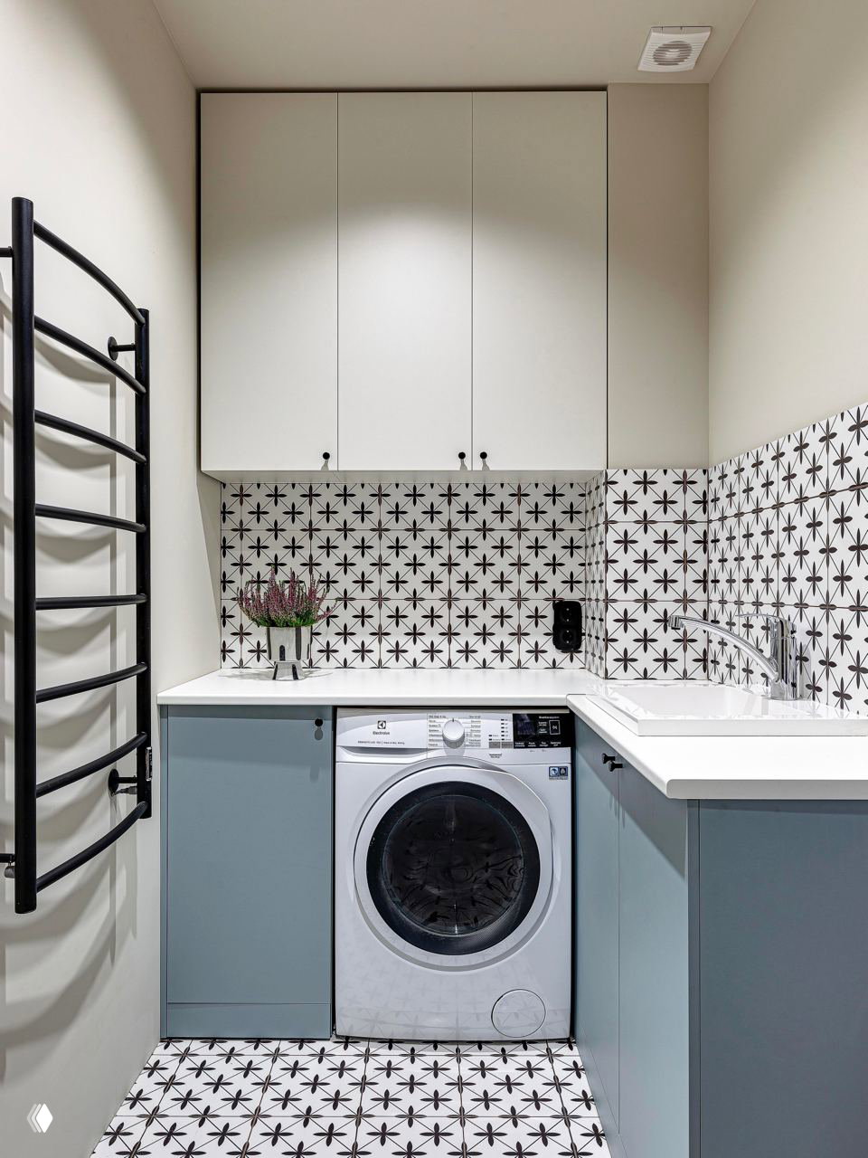 Практичная прачечная с встройкой: стиральная машина под столешницей, черная настенная сушилка-лесенка и декоративная плитка с орнаментом.