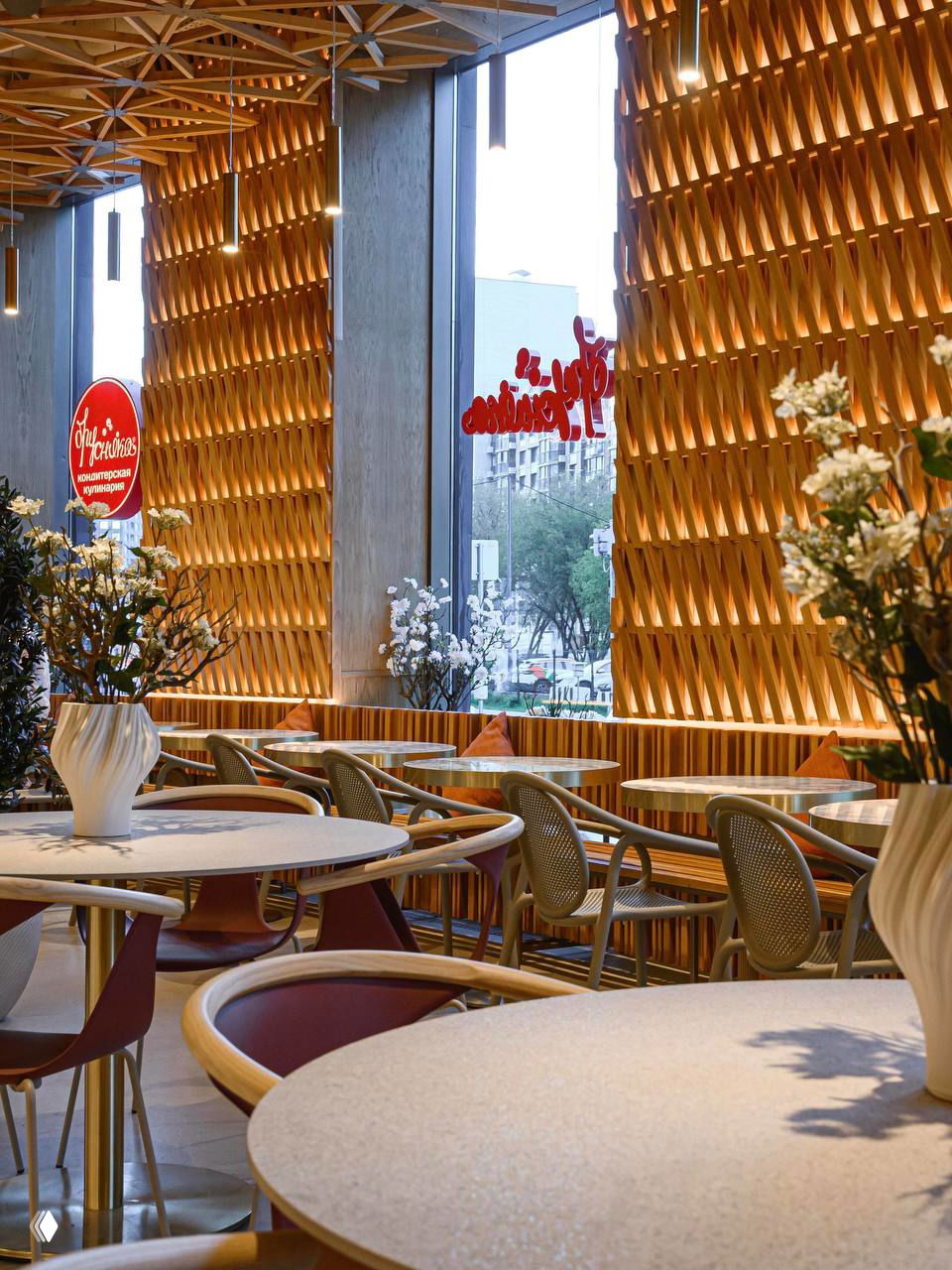 Зона гостей у высоких окон: сложное плетение из дерева на стене, круглые столы и итальянские стулья глубокого бордового оттенка.