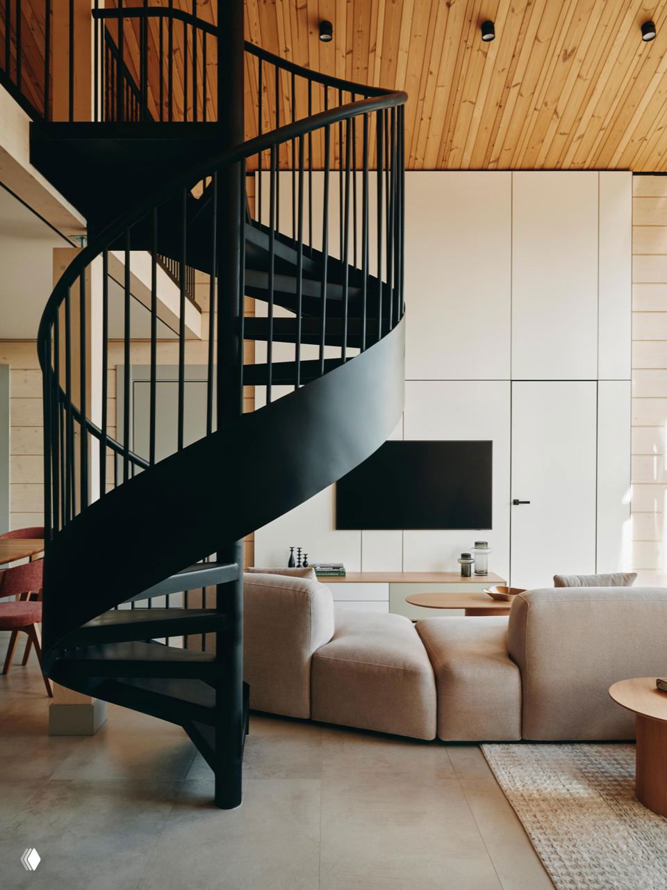 Крупный план чёрной металлической винтовой лестницы на фоне уютной гостиной с деревянным потолком, встроенной ТВ-зоной и светлыми панелями.