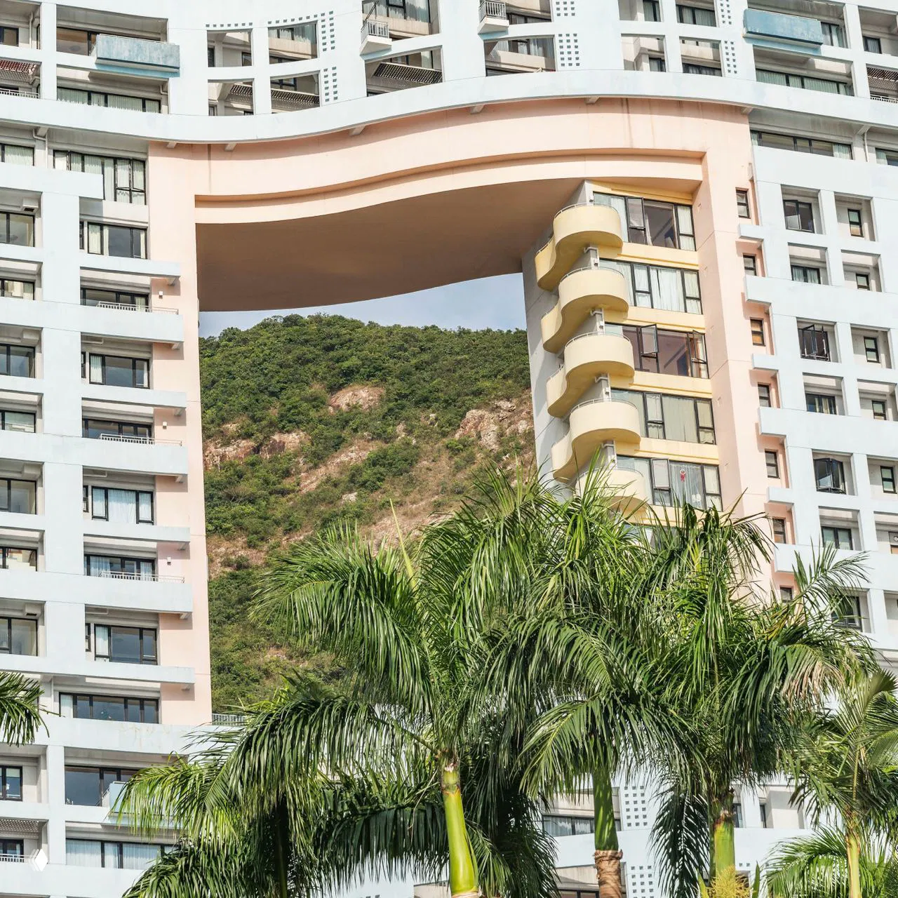 Крупный план архитектурного пролёта между секциями дома, пальмы на переднем плане и зелёные холмы в глубине создают городской ландшафт.