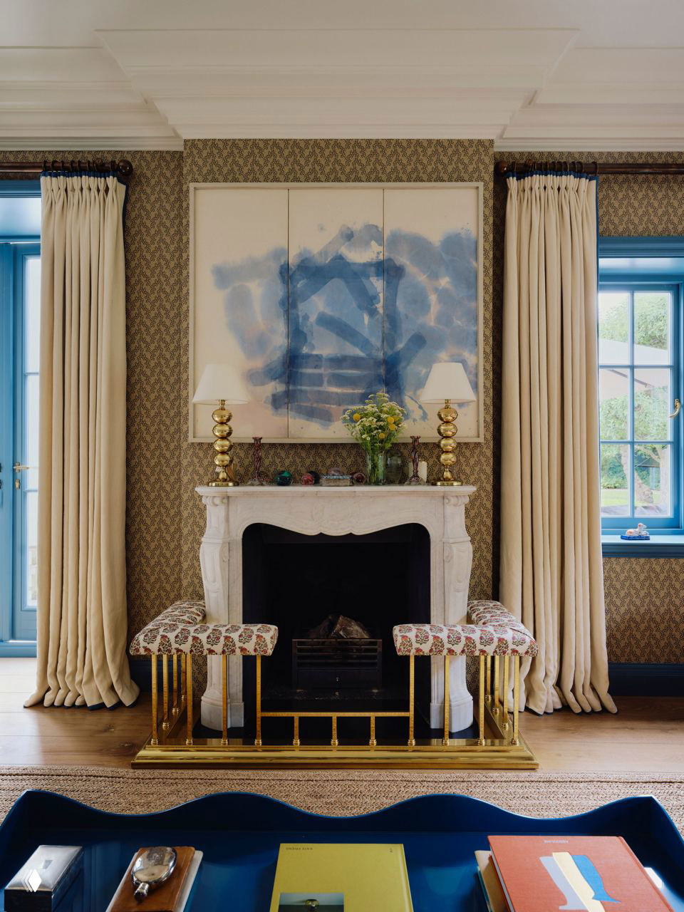 Классический камин в гостиной на фоне тёмных узорных обоев, две пуфы в передней части и кобальтовые оконные рамы по бокам для акцентов.