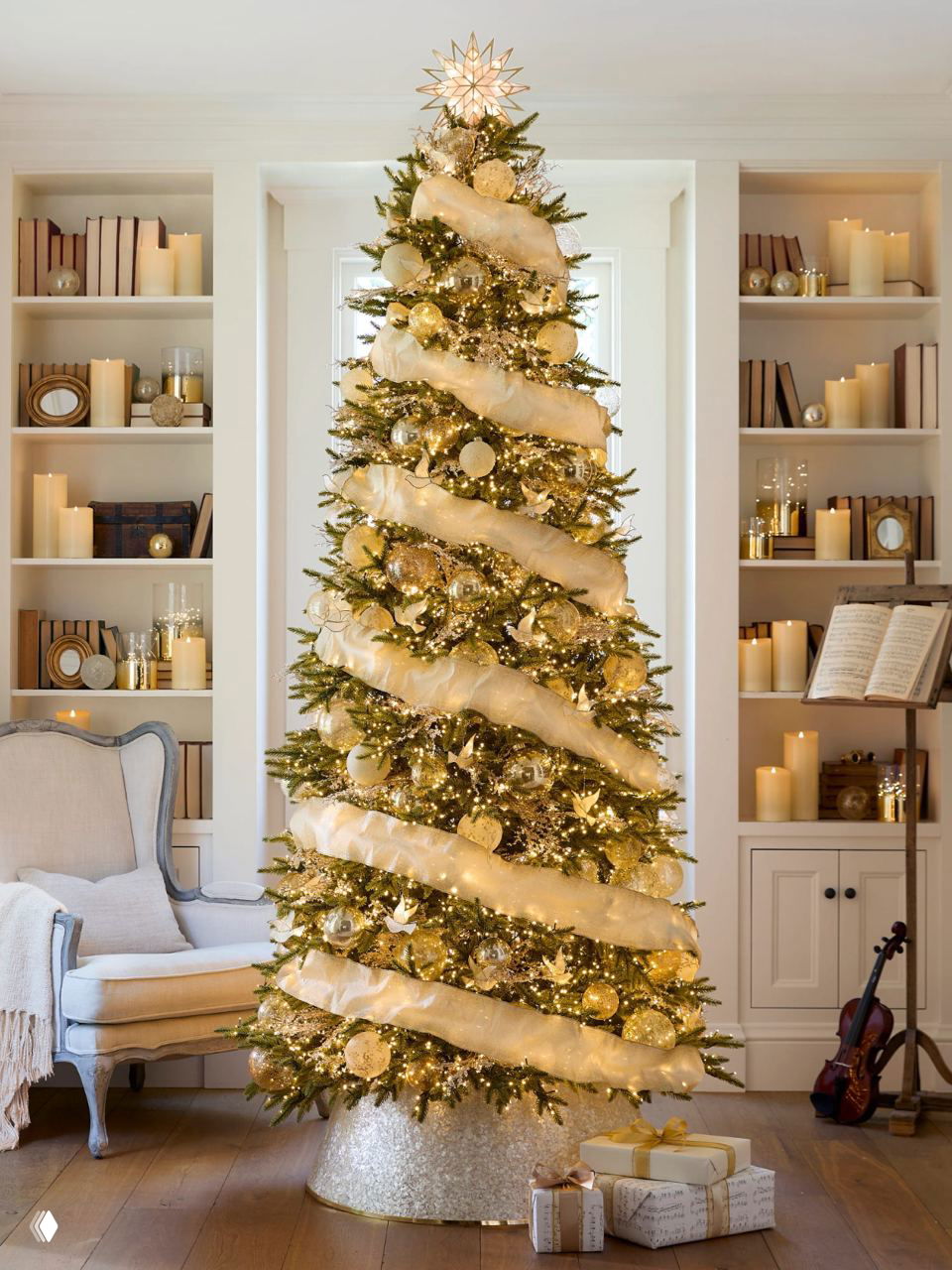 Классическая декоративная елка с широкой светлой тканевой гирляндой, золотыми шарами и множеством свечений на фоне книжных полок в гостиной.