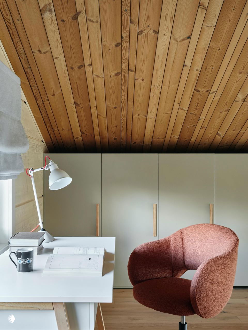 Рабочая ниша под скатом крыши: белый стол, настольная лампа, блокнот и коралловое кресло на фоне светлых встроенных шкафов и тёплого деревянного потолка.