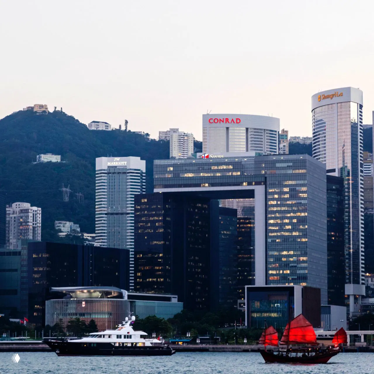 Деловой район у воды с коммерческими башнями, вывесками Conrad и JW Marriott на фоне холмов и причала, лодки у береговой линии.