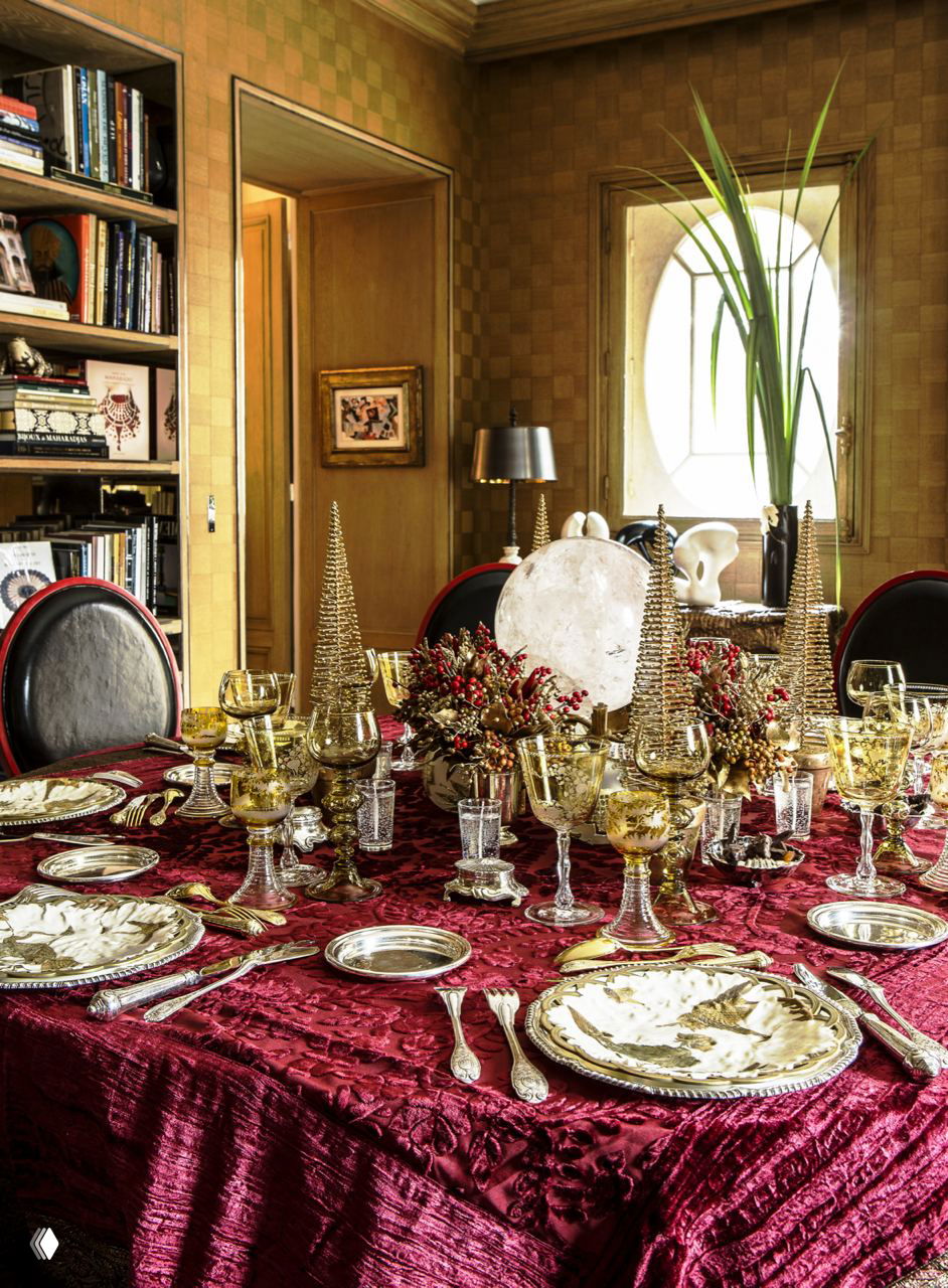 Праздничный сервированный стол с бордовой скатертью, золотой посудой, декоративными елочными фигурами и бокалами в интерьере столовой.