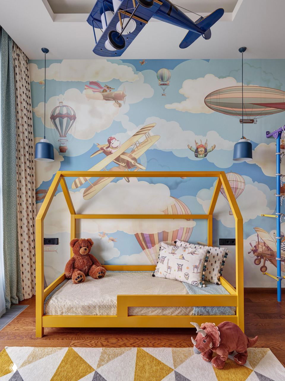 Детская с домиком-кроватью жёлтого цвета на деревянном полу, обоями с иллюстрациями самолётов и воздушных шаров и мягкими игрушками у кровати для игровой атмосферы.