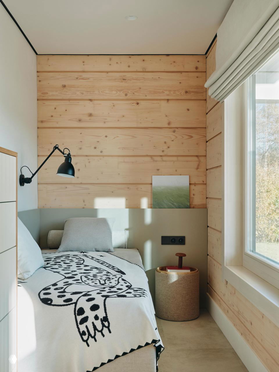 Небольшая спальня в доме из клееного бруса: кровать у окна, деревянные панели стен, декоративный текстиль с рисунком и мягкое дневное освещение.