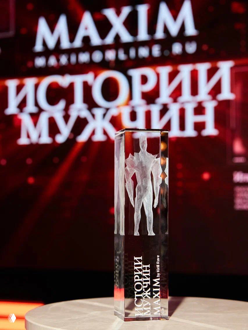 Приз проекта «Истории мужчин MAXIM» на переднем плане на фоне размытого экрана с логотипом MAXIM — награда в стеклянном блоке с гравировкой