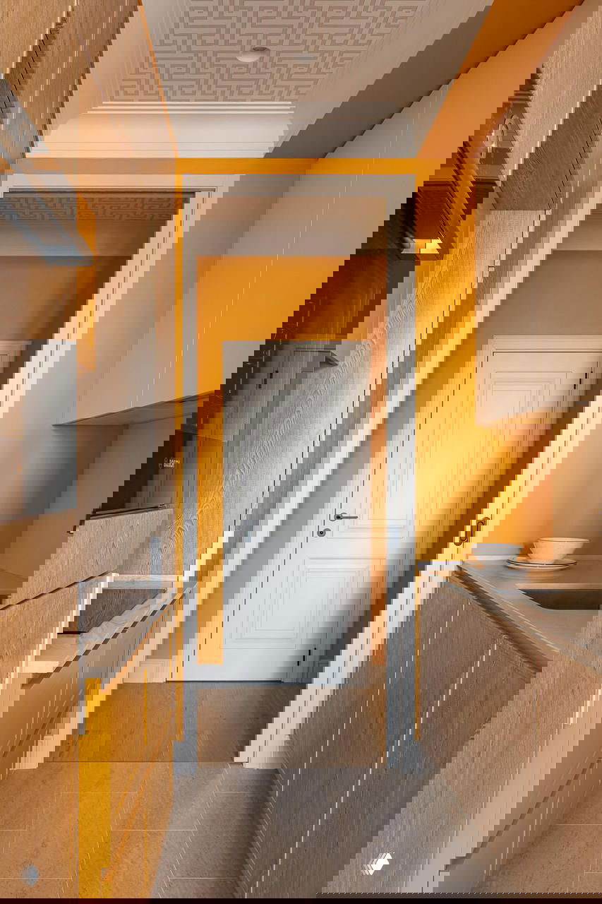 Вытянутая кухня с ярко-оранжевыми акцентами и деревянными фасадами, проход между рабочими зонами и светлая дверь в торце — пример использования цвета в узком помещении.