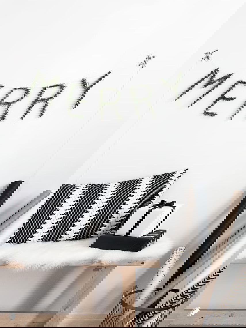 Минималистичная белая стена с надписью MERRY из хвойных веточек; скамья с черно‑белой подушкой, подарки в лаконичной упаковке и окружение ручной работы.