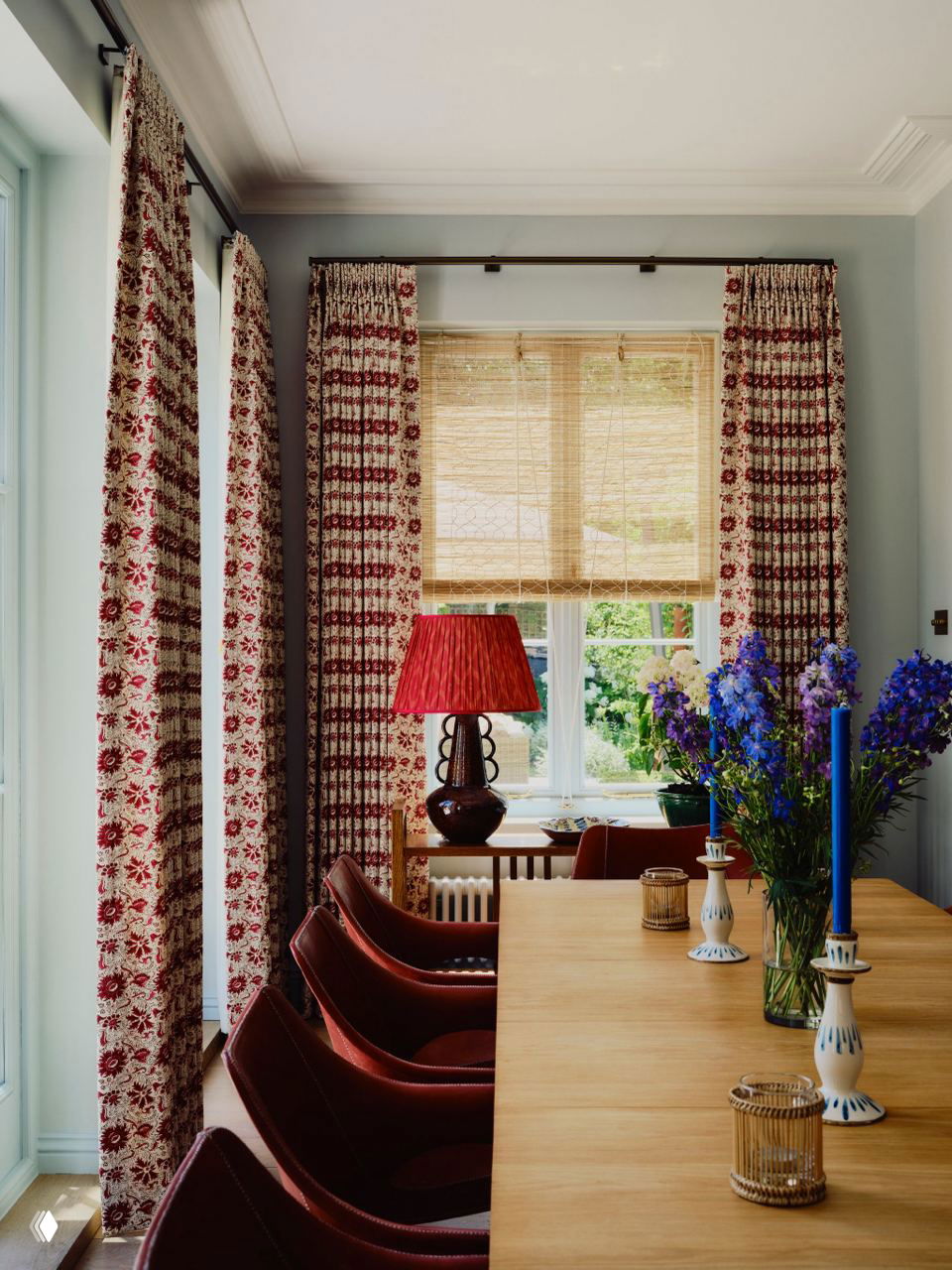 Обеденная зона с длинным деревянным столом, бордовыми стульями, высокими шторами с узором и декоративными предметами на столе у окна.
