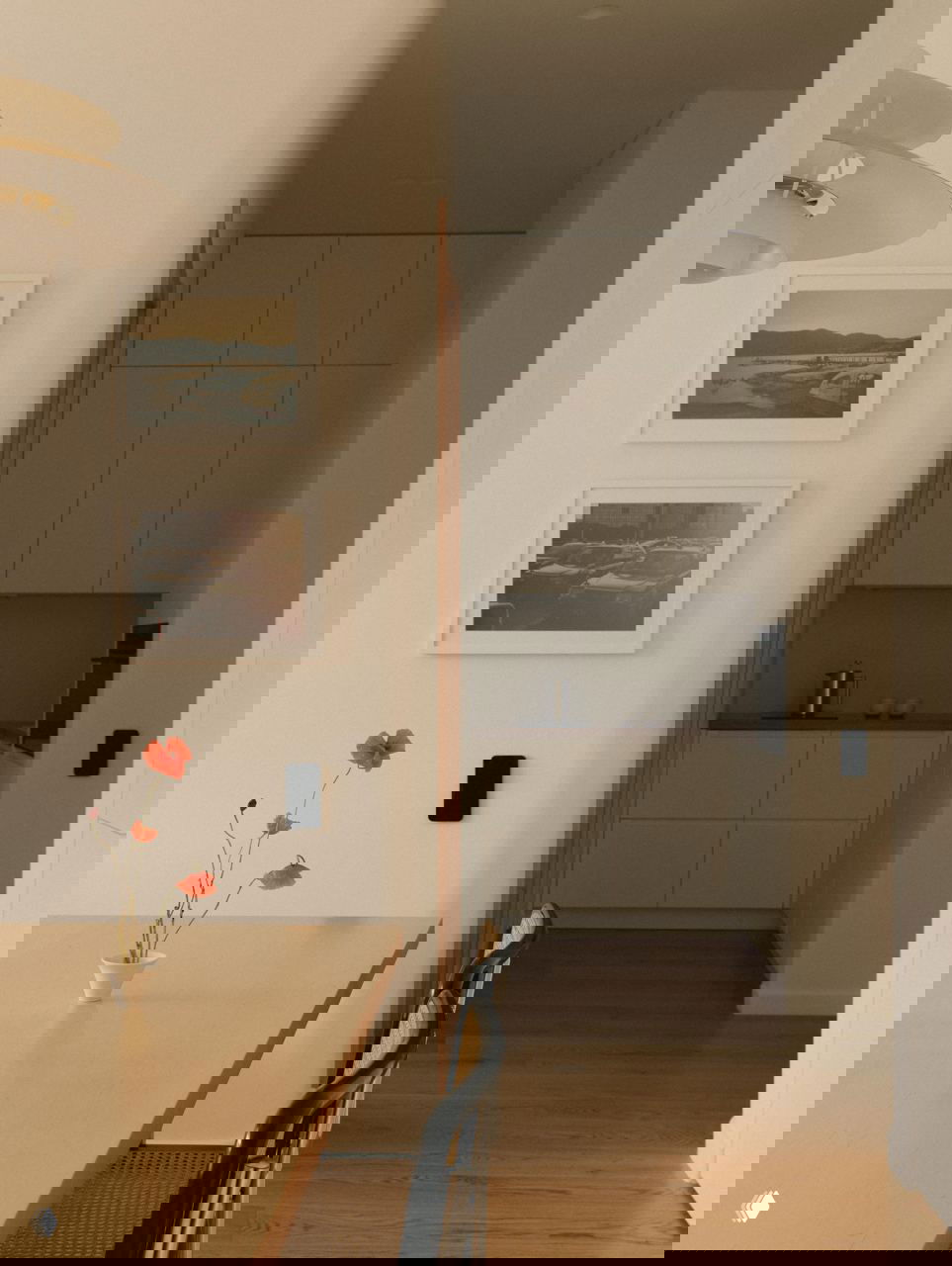 Столовая и проход к кухне: деревянный стол с вазой и цветком, простые стулья и минималистичная кухня в глубине, натуральные материалы и спокойная палитра.