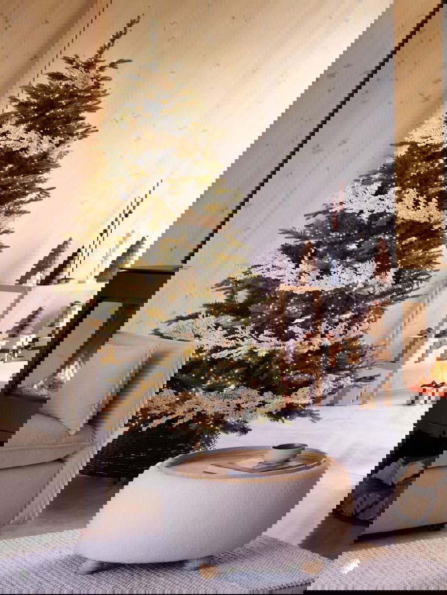 Классическая рождественская сцена с высокой елкой, украшенной мелкими огнями и белыми декоративными веточками, рядом кресло, ковер и теплый камин.