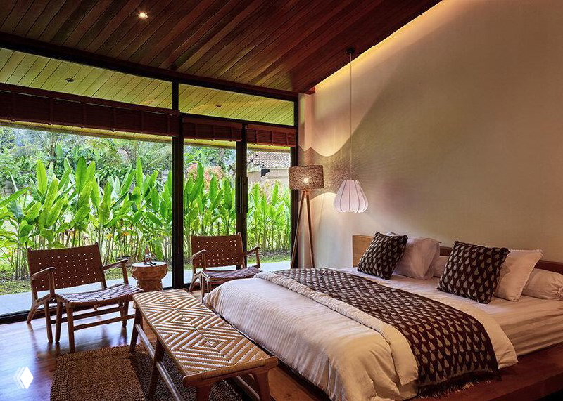 Интерьерная фотография спальни с деревянным потолком и большими проёмами, где видна связь внутреннего пространства с зеленью за пределами.