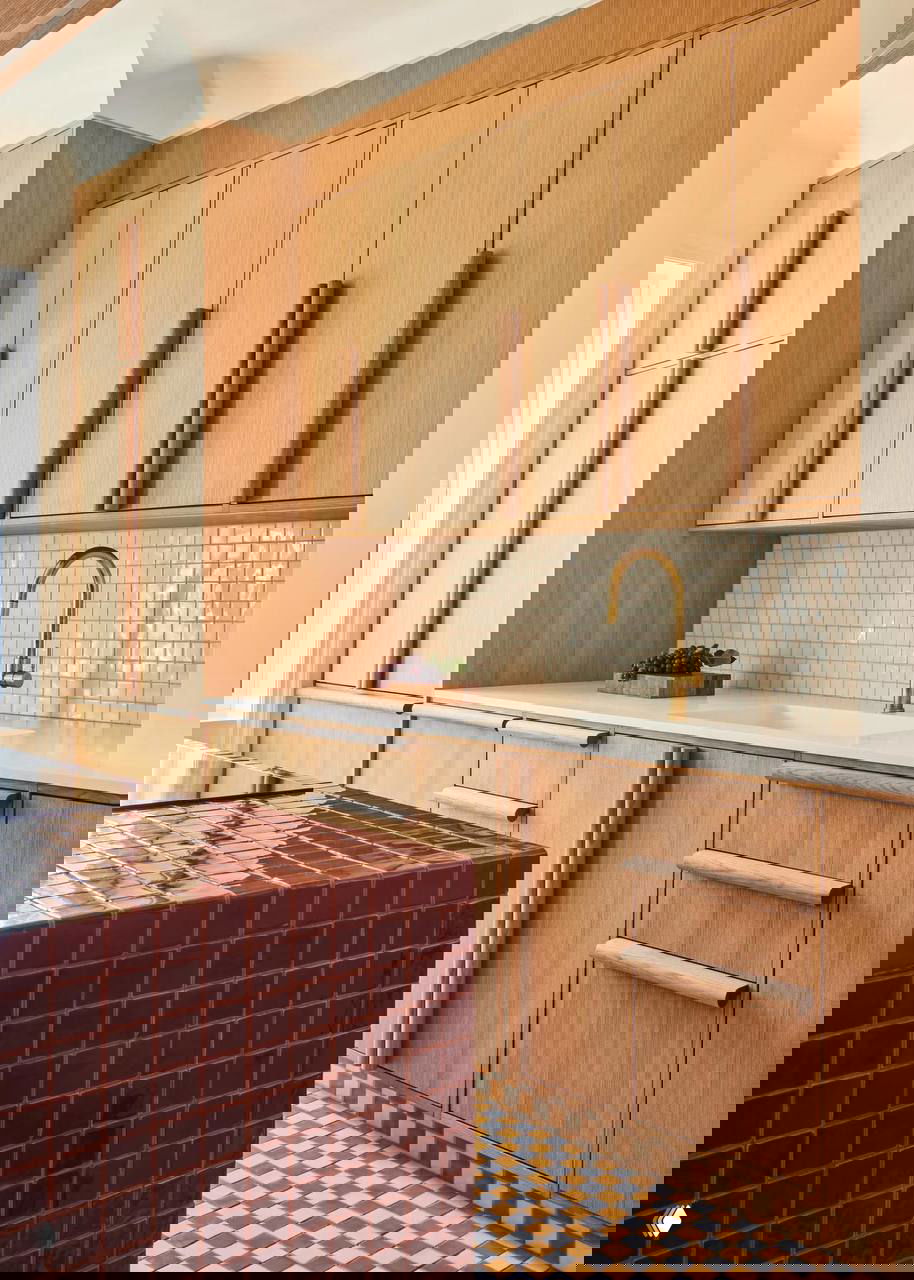 Тёплая деревянная параллельная кухня с модулями под дерево, белой столешницей и центральным островком, а также декоративной красной плиткой на переднем плане.