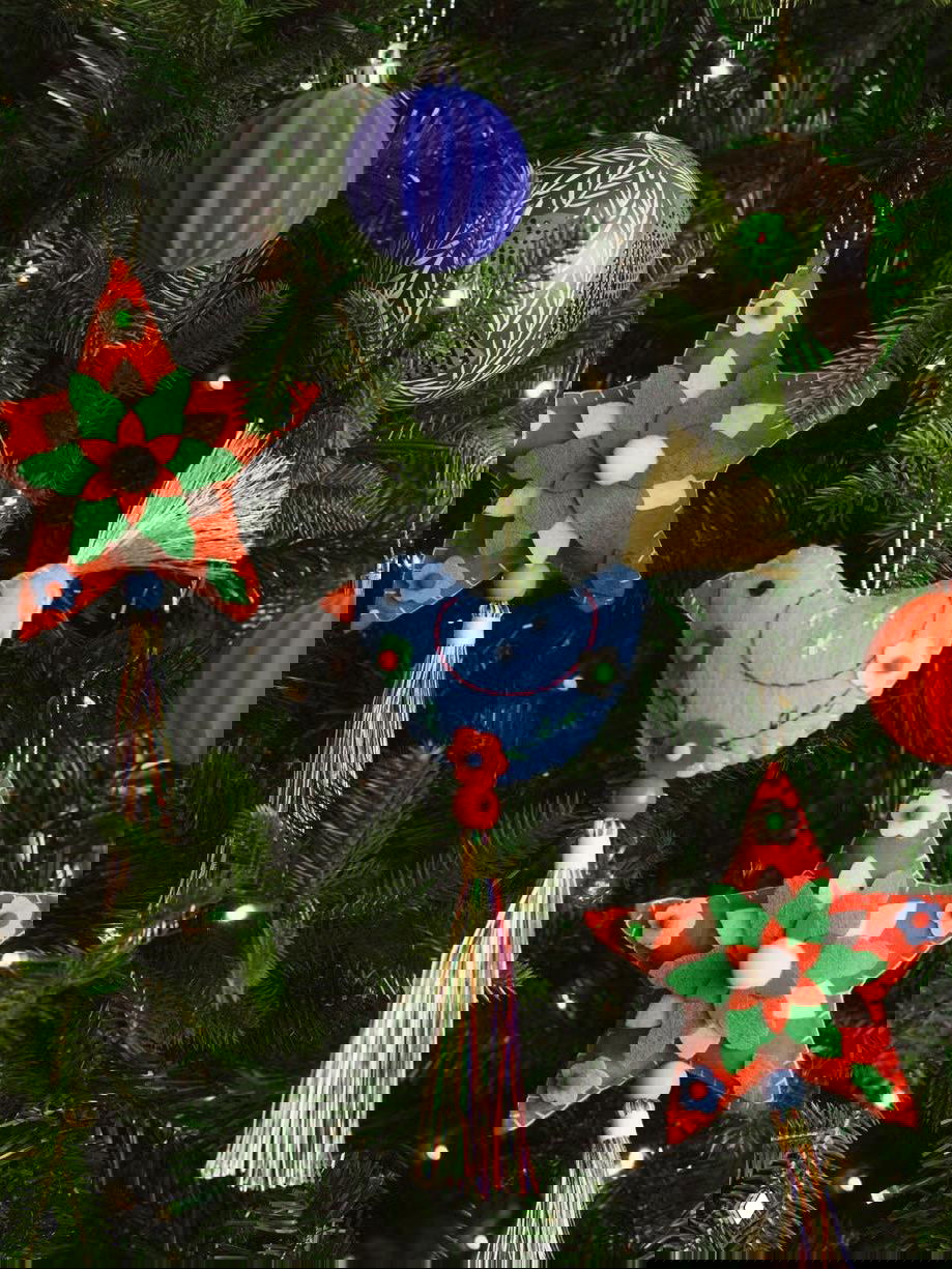 Крупный план текстильных и фетровых елочных игрушек: птички, звезды и кисточки, ручная вышивка и разнообразные цвета на фоне зелени ветвей.