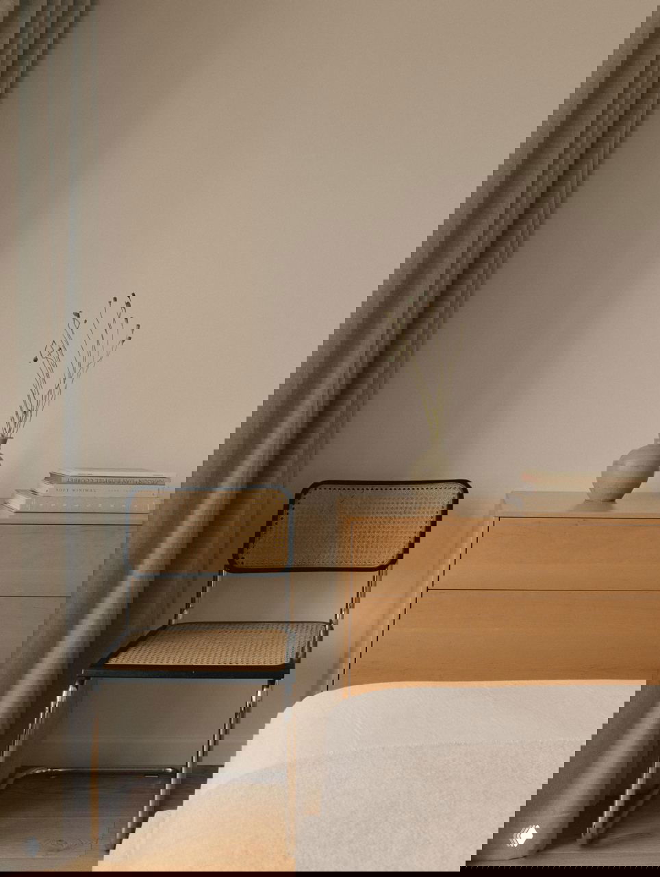 Уголок интерьера: плетёный стул у деревянного комода, керамическая ваза с сухоцветами и книги, акцент на естественных материалах и минимализме.