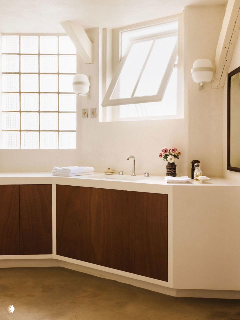 Светлая ванная зона с угловой столешницей и деревянными фасадами шкафчиков, большое окно пропускает мягкий дневной свет в интерьер.